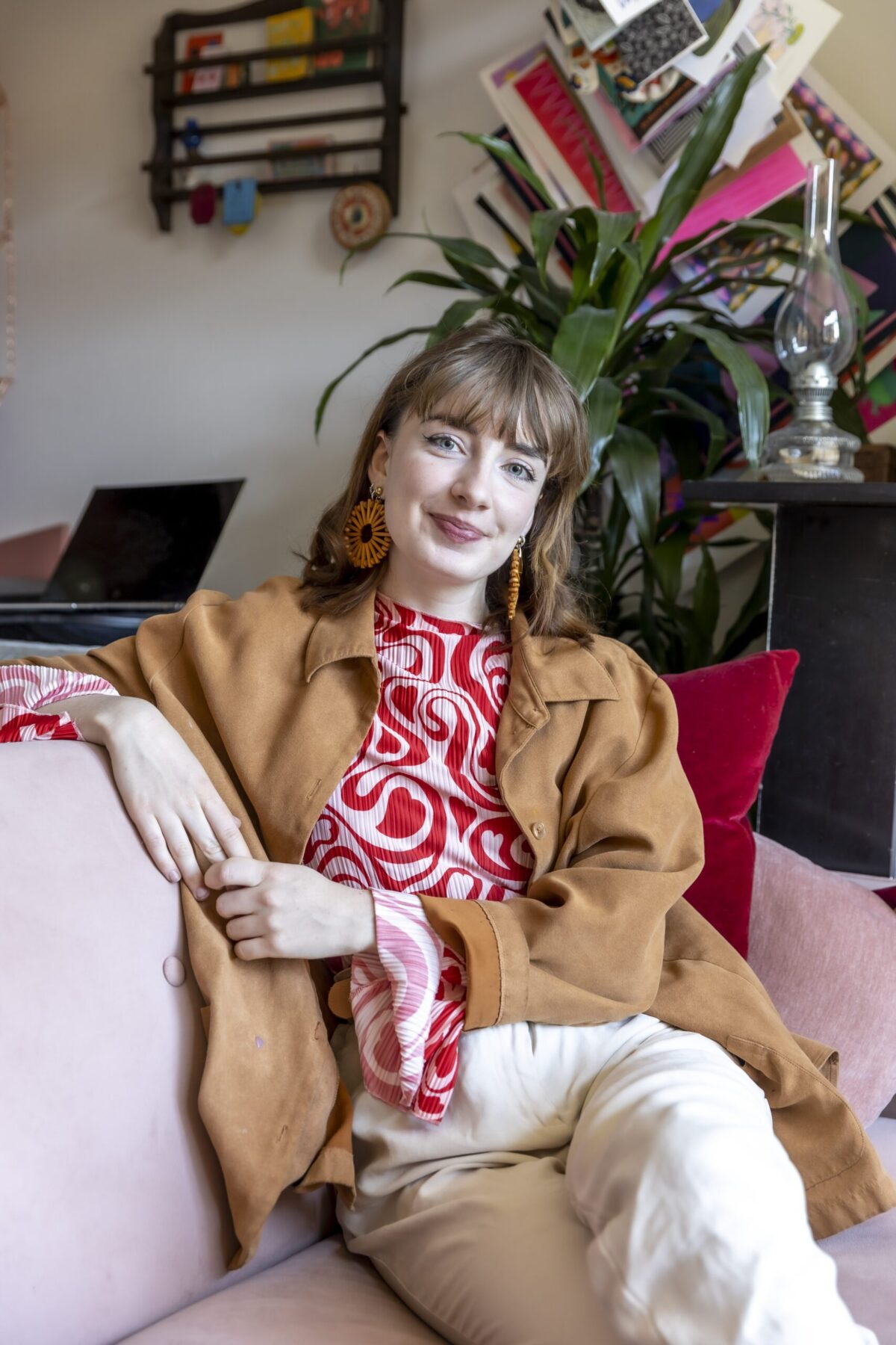 A colorful riso print illustrator and motion designer from Utrecht lounges on a pink couch, their long hair cascading over a brown jacket and red patterned top. Smiling warmly, they rest one arm on the couch amid a backdrop of plants and eclectic wall decorations.
